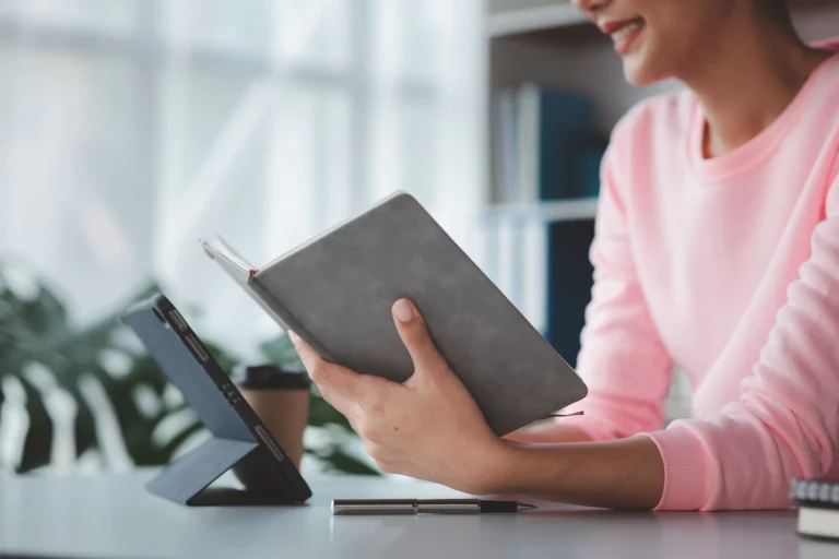 Mulher segurando um caderno cinza em uma mesa com tablet e café.